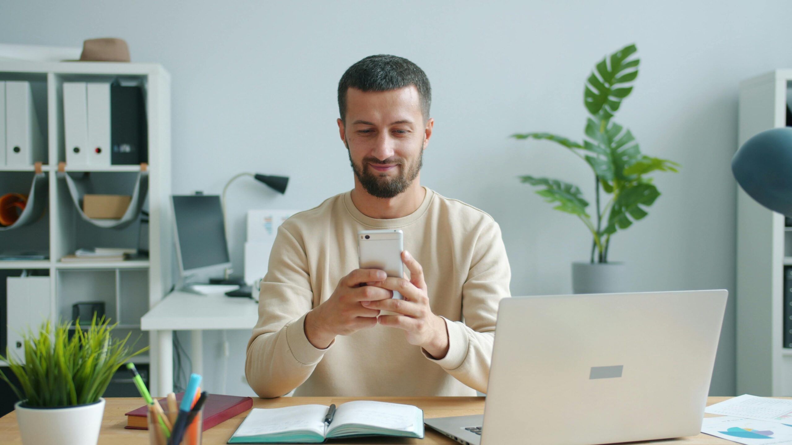 A man sitting at a desk looks at his smartphone, surrounded by office supplies in a bright modern office.